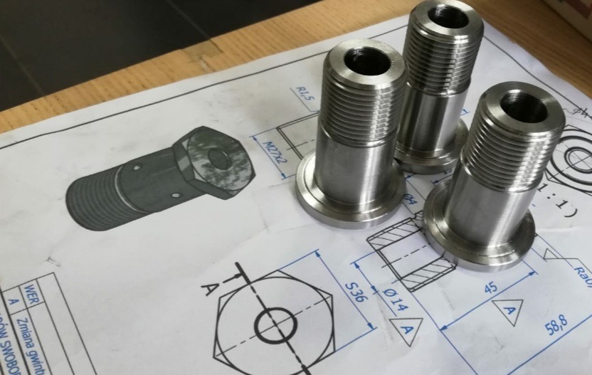 Trzy metalowe elementy z gwintem stojące na technicznym rysunku z wymiarami. Precyzyjna obróbka metalu, detal wykonany z dbałością o szczegóły.