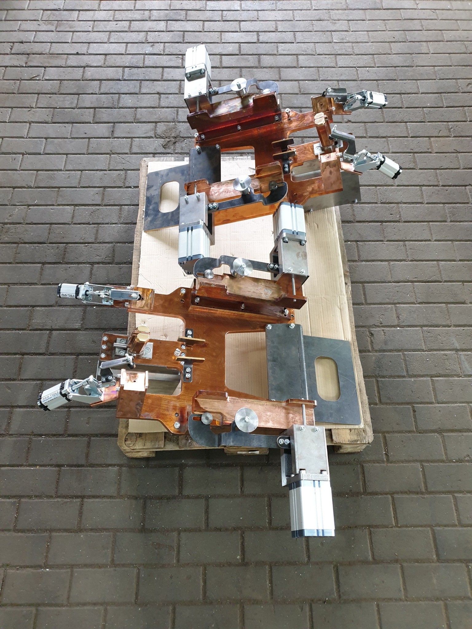 Precyzyjnie wykonany, miedziano-stalowy uchwyt spawalniczy na palecie, z widocznymi elementami pneumatycznymi i mocowaniami, na tle szarej, brukowej podłogi.