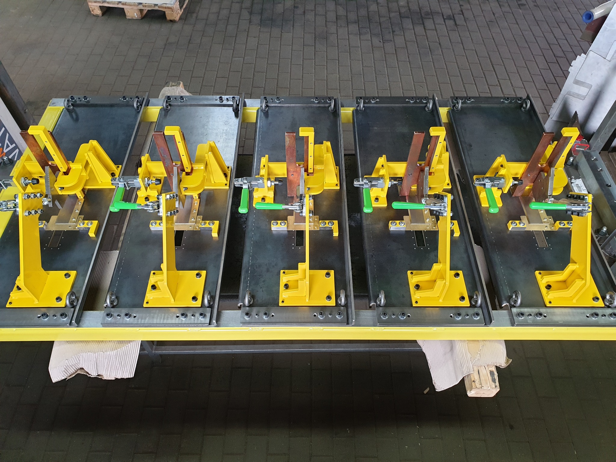 Precyzyjne, żółte oprzyrządowanie spawalnicze na szarych płytach, ułożone rzędem na konstrukcji. Widoczne elementy mocujące i regulacyjne z zielonymi uchwytami.
