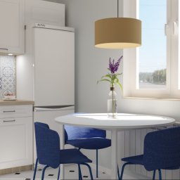 Joanna Staniszewska - Jasna kuchnia z białymi szafkami, wzorzystą mozaiką nad blatem, okrągłym stołem i niebieskimi krzesłami. Na parapecie doniczki z ziołami, nad stołem lampa.