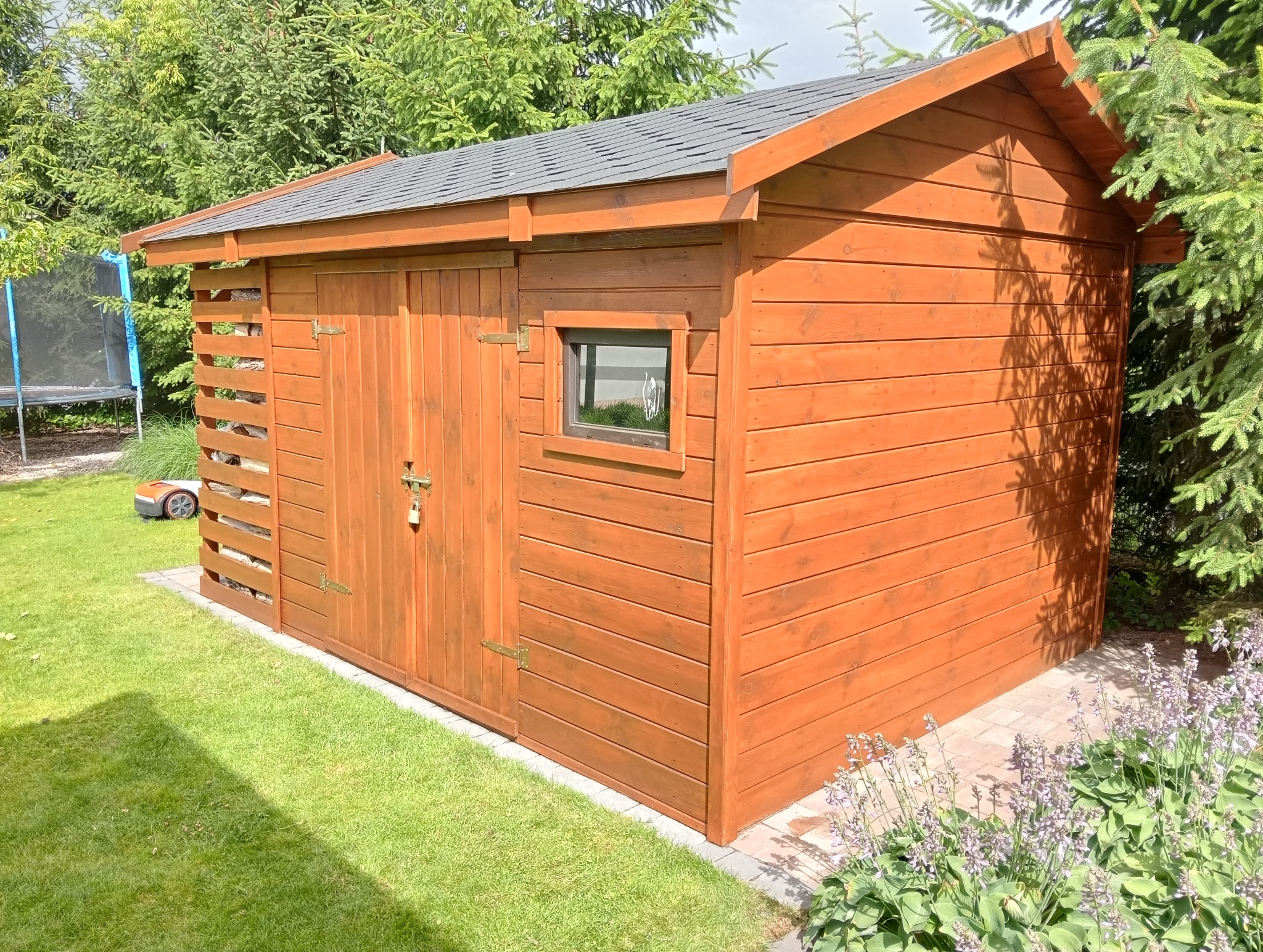 Drewniana szopa ogrodowa z szarym dachem, widoczna z boku na tle zielonego trawnika i drzew. Część ściany ażurowa, odsłaniająca składowane drewno.