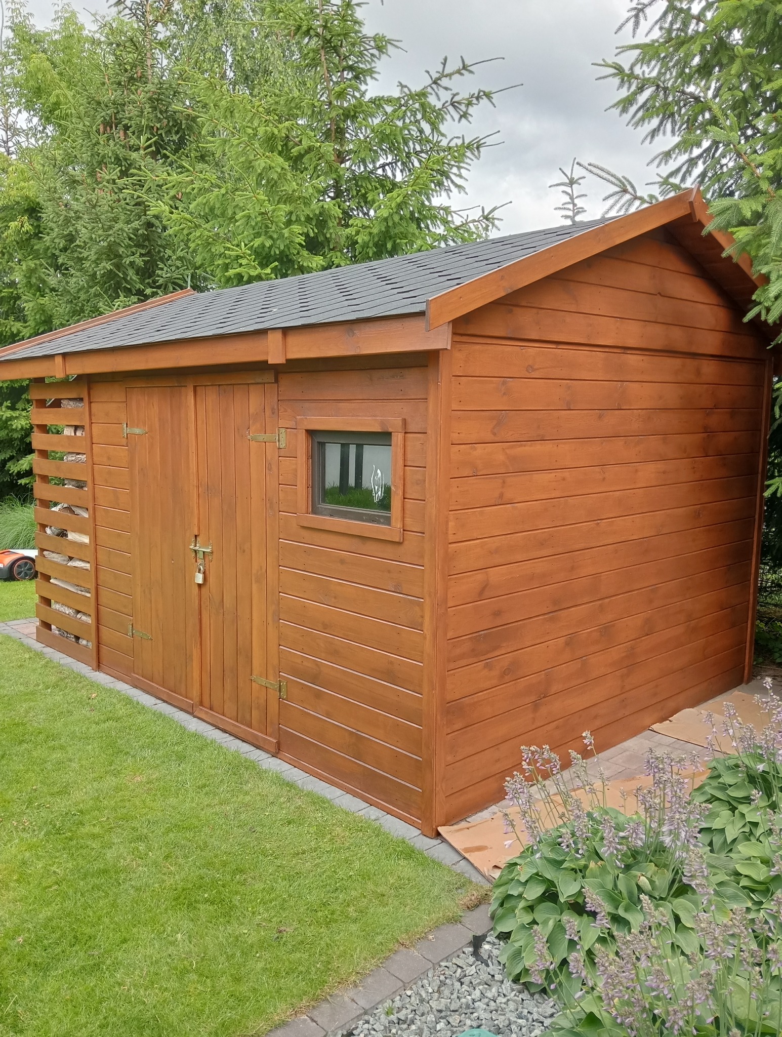 Drewniana, brązowa szopa ogrodowa z oknem i dachem krytym gontem bitumicznym, otoczona zielenią i kwiatami. Świeżo postawiona konstrukcja na trawie.