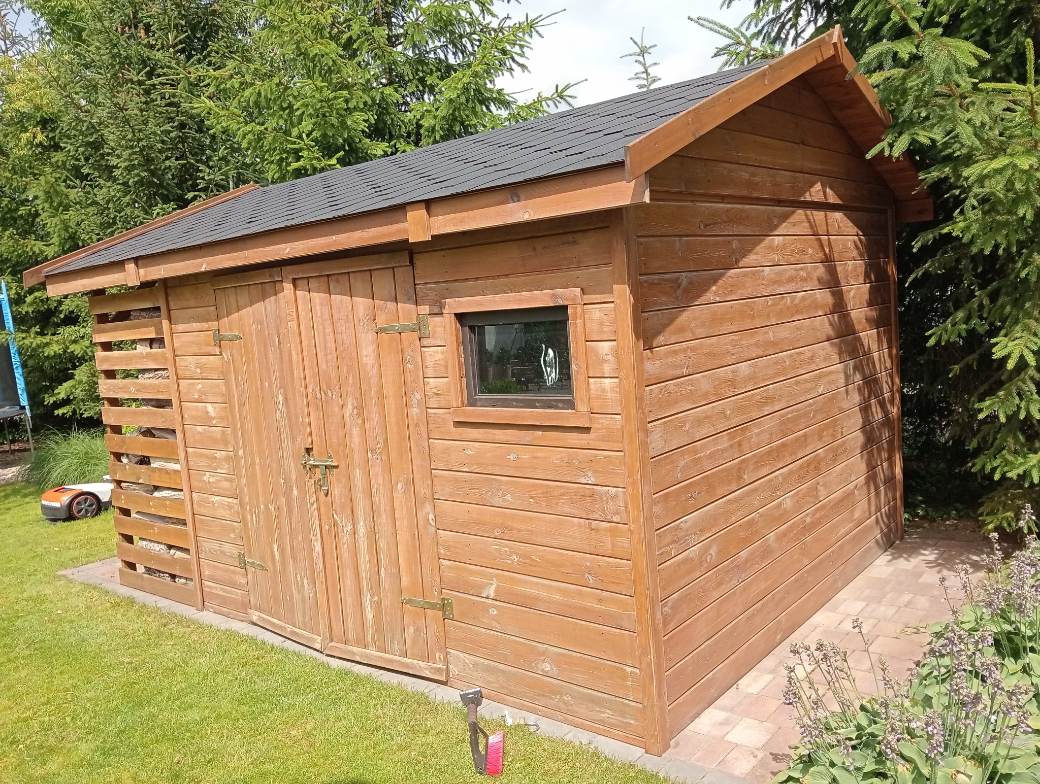 Drewniana szopa ogrodowa z czarnym, spadzistym dachem, oknem i drzwiami na posesji z zieloną trawą i drzewami iglastymi w tle. Widoczny robot koszący.