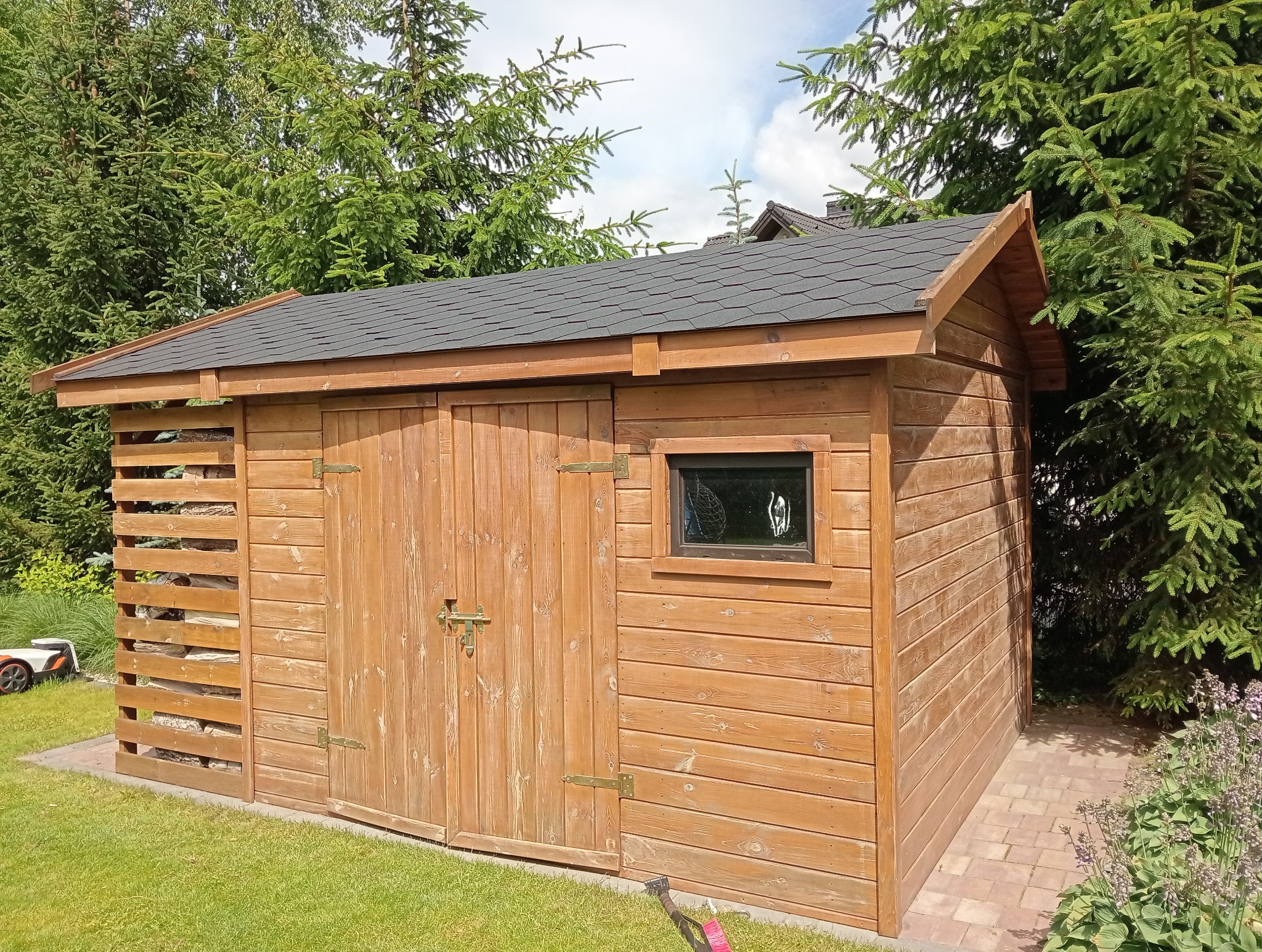 Drewniana szopa ogrodowa z czarnym dachem na tle zieleni, widoczna z bliska, z elementami wyposażenia wewnątrz i na zewnątrz.