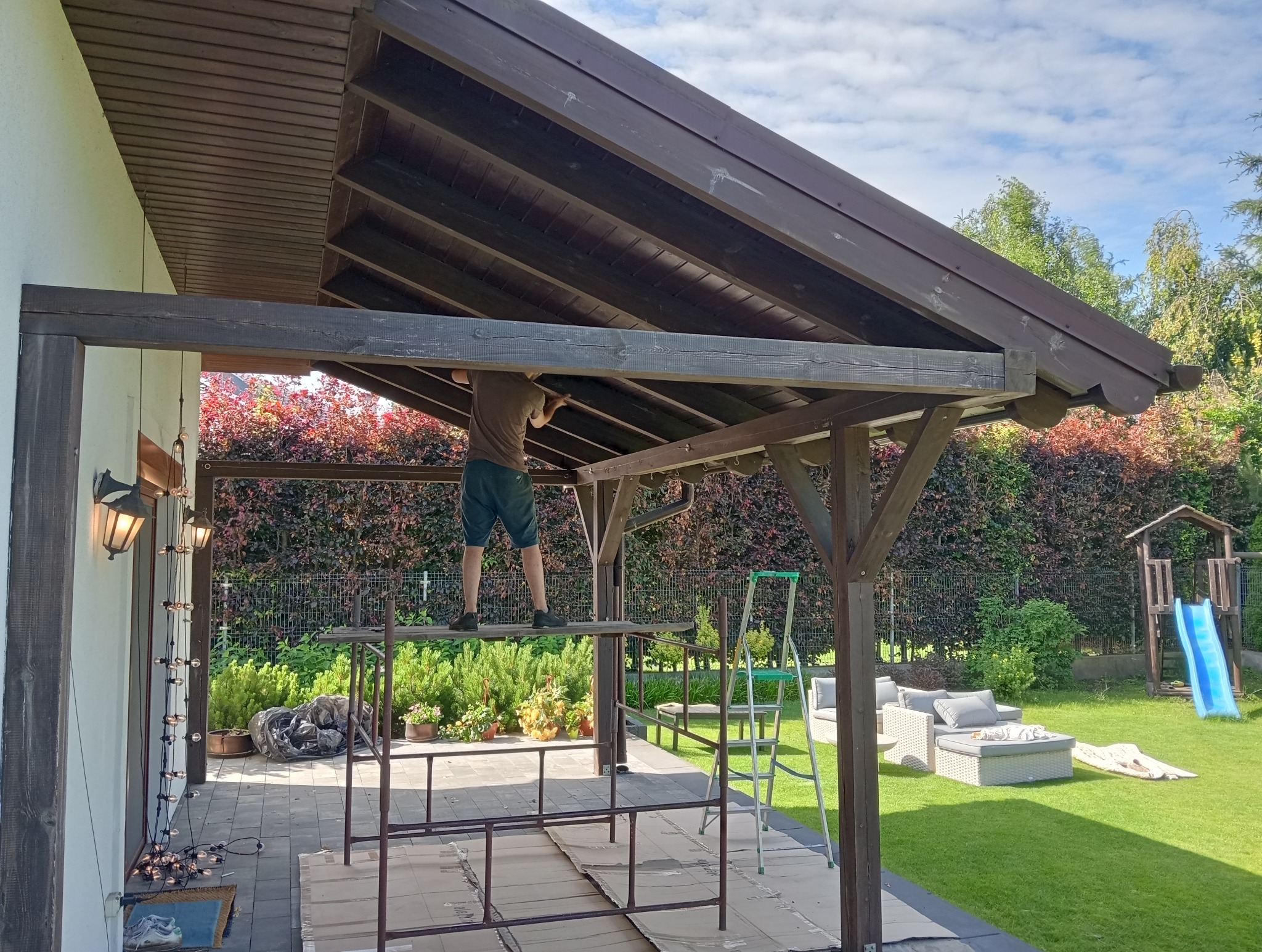 Montaż drewnianej konstrukcji zadaszenia tarasu przez mężczyznę na rusztowaniu, widoczny fragment ogrodu z meblami wypoczynkowymi i placem zabaw.