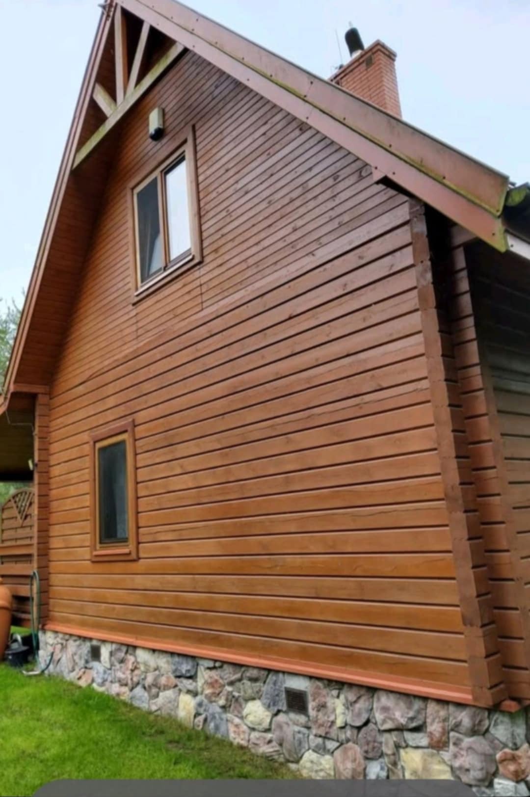 Dom z brązową, drewnianą elewacją panelową i kamienną podmurówką. Widoczne dwa okna w drewnianych ramach i fragment dachu. Ujęcie z dołu, lekko z prawej strony.