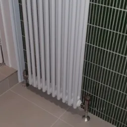 Biały grzejnik żeberkowy w łazience z zieloną mozaiką na ścianie i beżowymi płytkami na podłodze. Widoczne zawory termostatyczne.