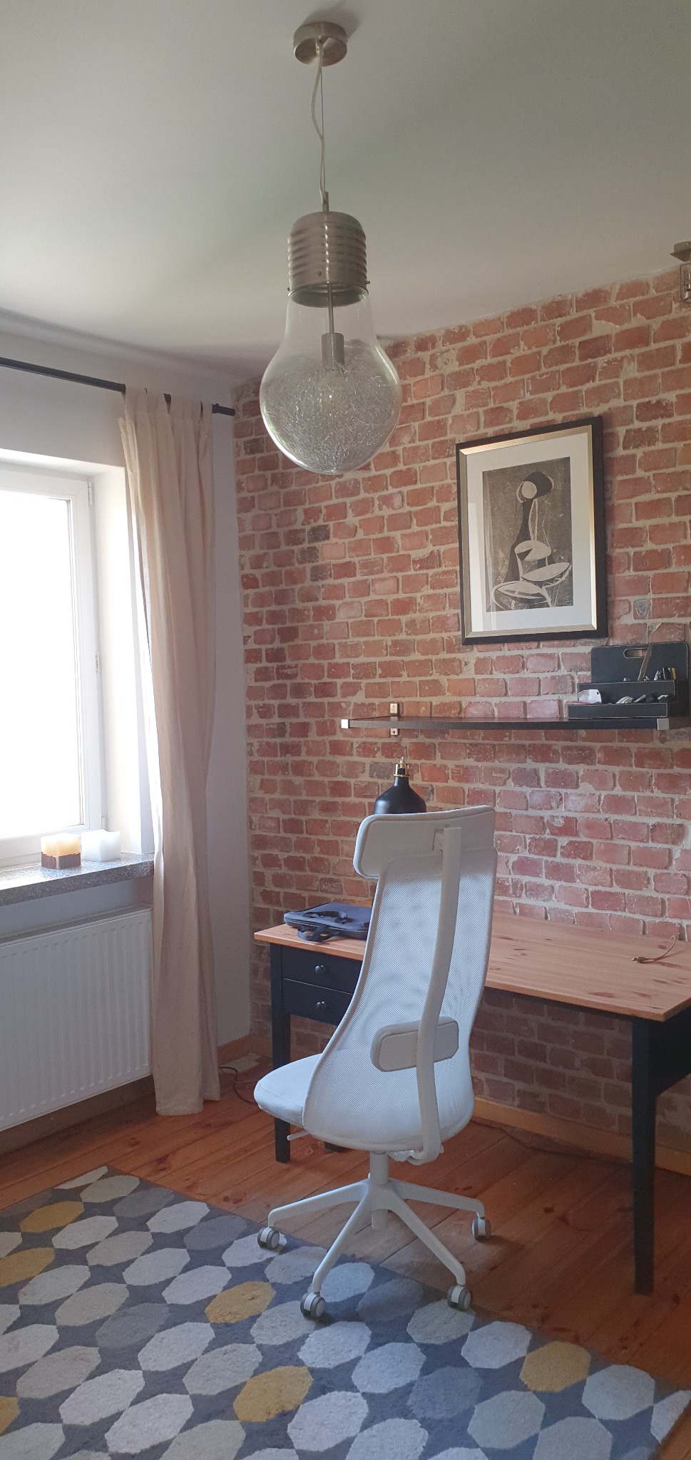 Pokój z ceglaną ścianą i biurkiem, oświetlony lampą w kształcie żarówki. Białe krzesło biurowe na tle wzorzystego dywanu. Nowoczesne wykończenie wnętrza.
