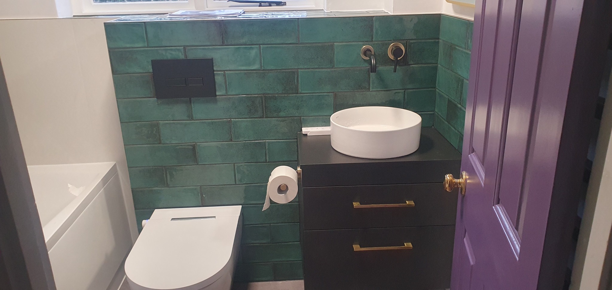 Wykończona łazienka: turkusowa glazura, czarna szafka z umywalką i złotymi uchwytami, wisząca toaleta z deską myjącą i fioletowe drzwi.