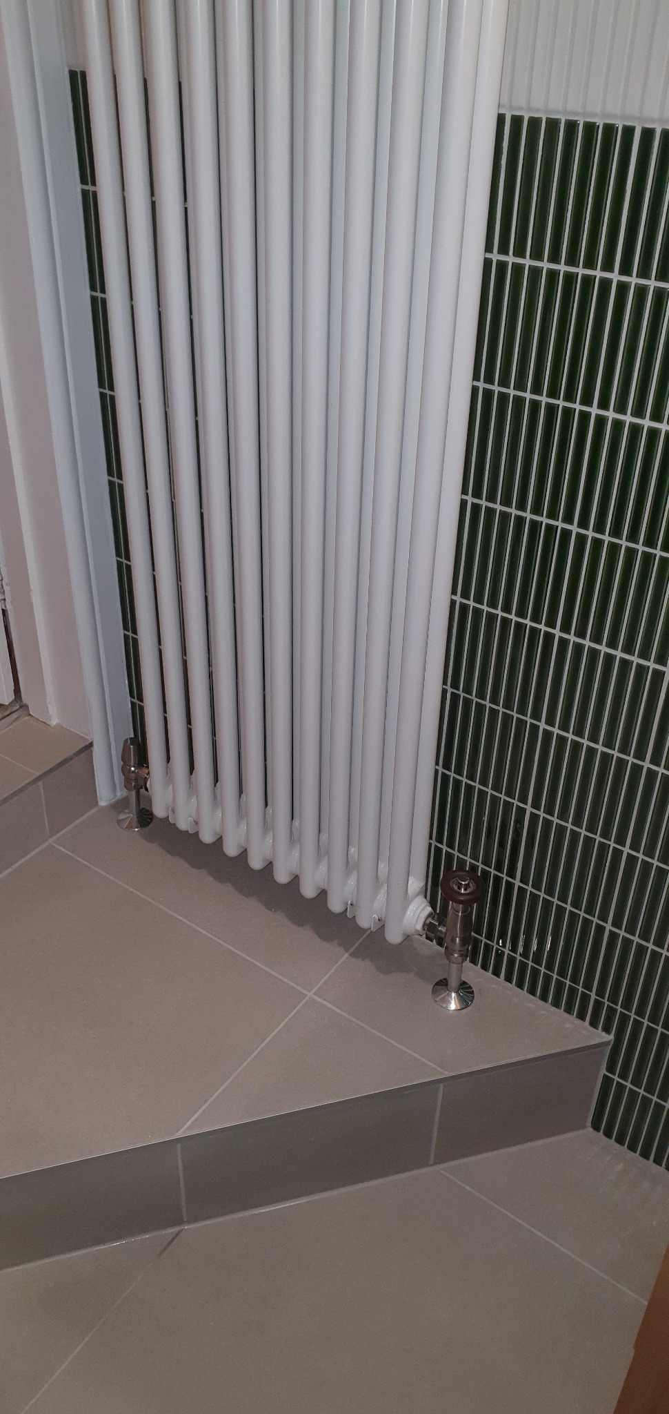 Biały grzejnik żeberkowy w łazience z zieloną mozaiką na ścianie i beżowymi płytkami na podłodze. Widoczne zawory termostatyczne.