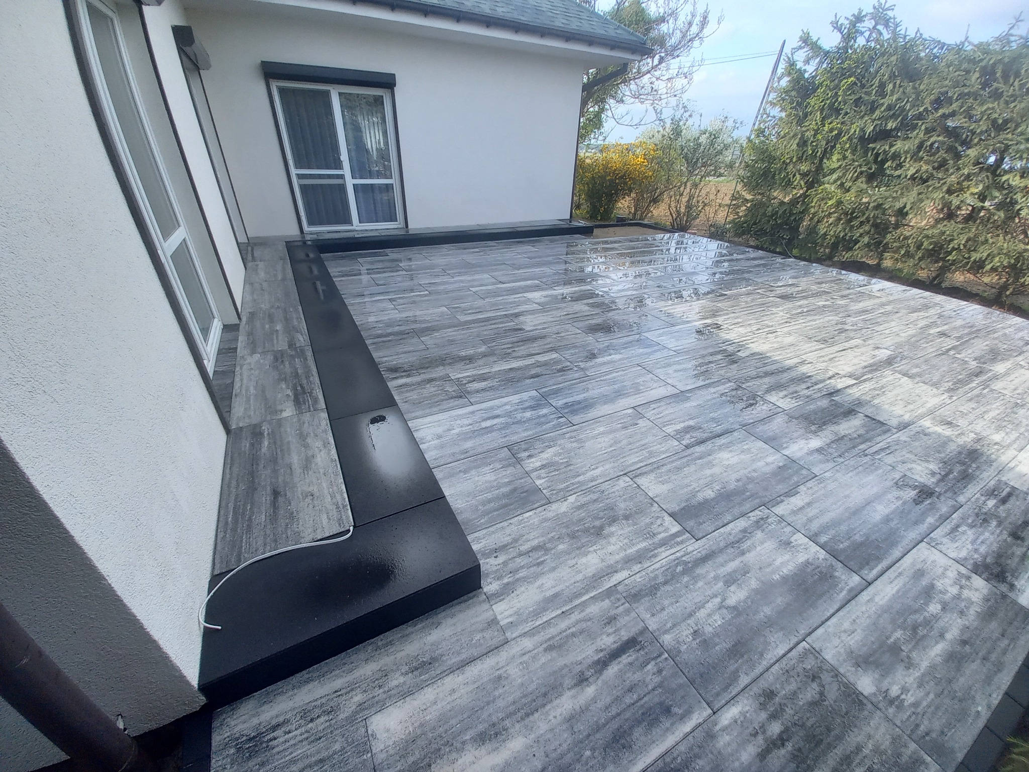 Szara kostka brukowa ułożona przy domu, z czarnym obrzeżem i odpływem liniowym. Widoczne rośliny w tle i fragment elewacji budynku.