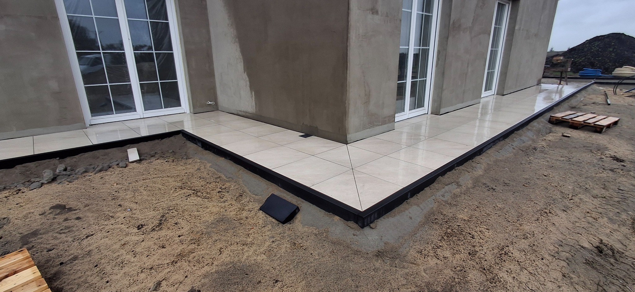 Jasne płytki tarasowe ułożone przy budynku z białymi oknami, obrzeża w czarnych profilach. Widoczny fragment terenu przygotowanego pod dalsze prace.