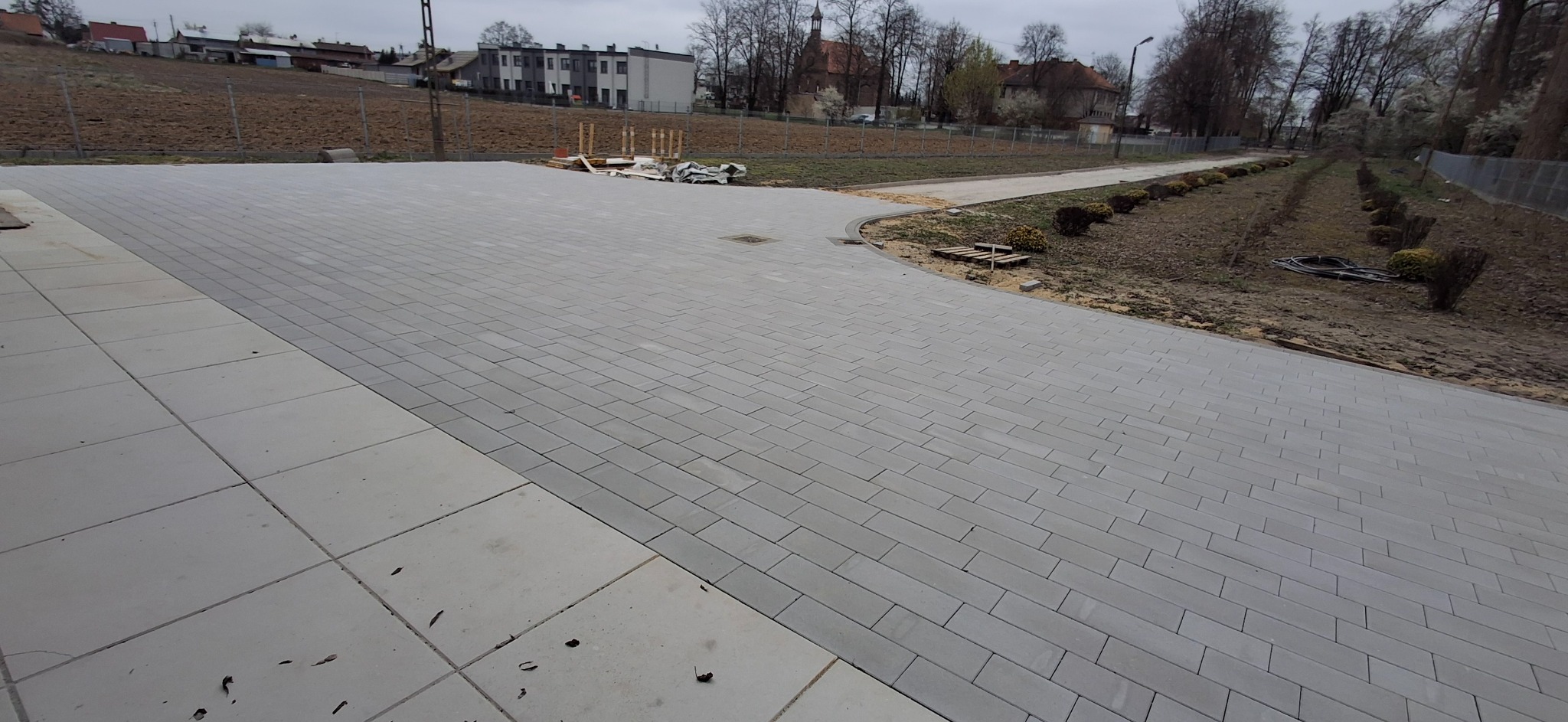 Nowa, szara kostka brukowa ułożona na posesji, widok z góry. W tle budynki mieszkalne i drzewa. Częściowo widoczne palety i narzędzia.