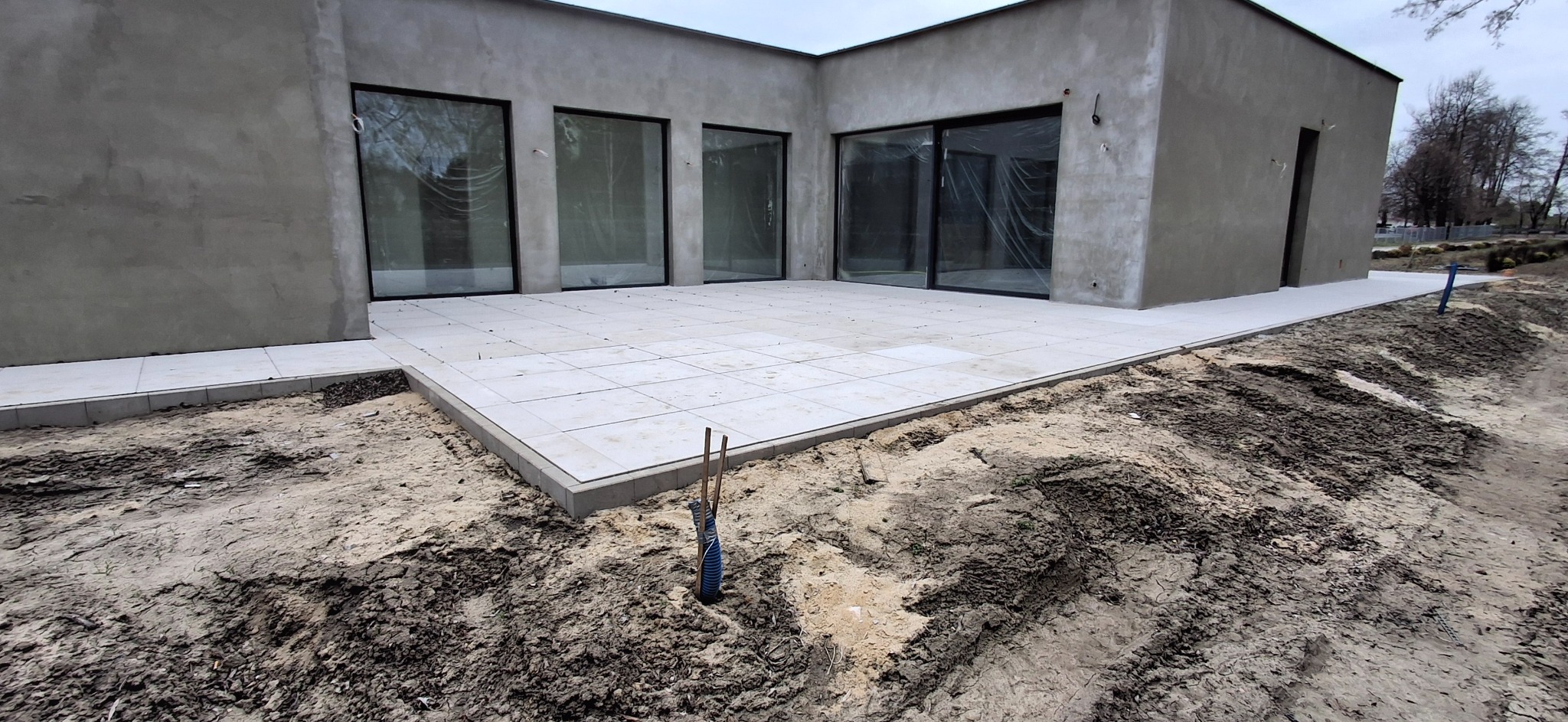 Jasna, kwadratowa kostka brukowa na tarasie nowoczesnego domu z dużymi oknami. W tle widoczny teren przygotowany pod dalsze prace ziemne.