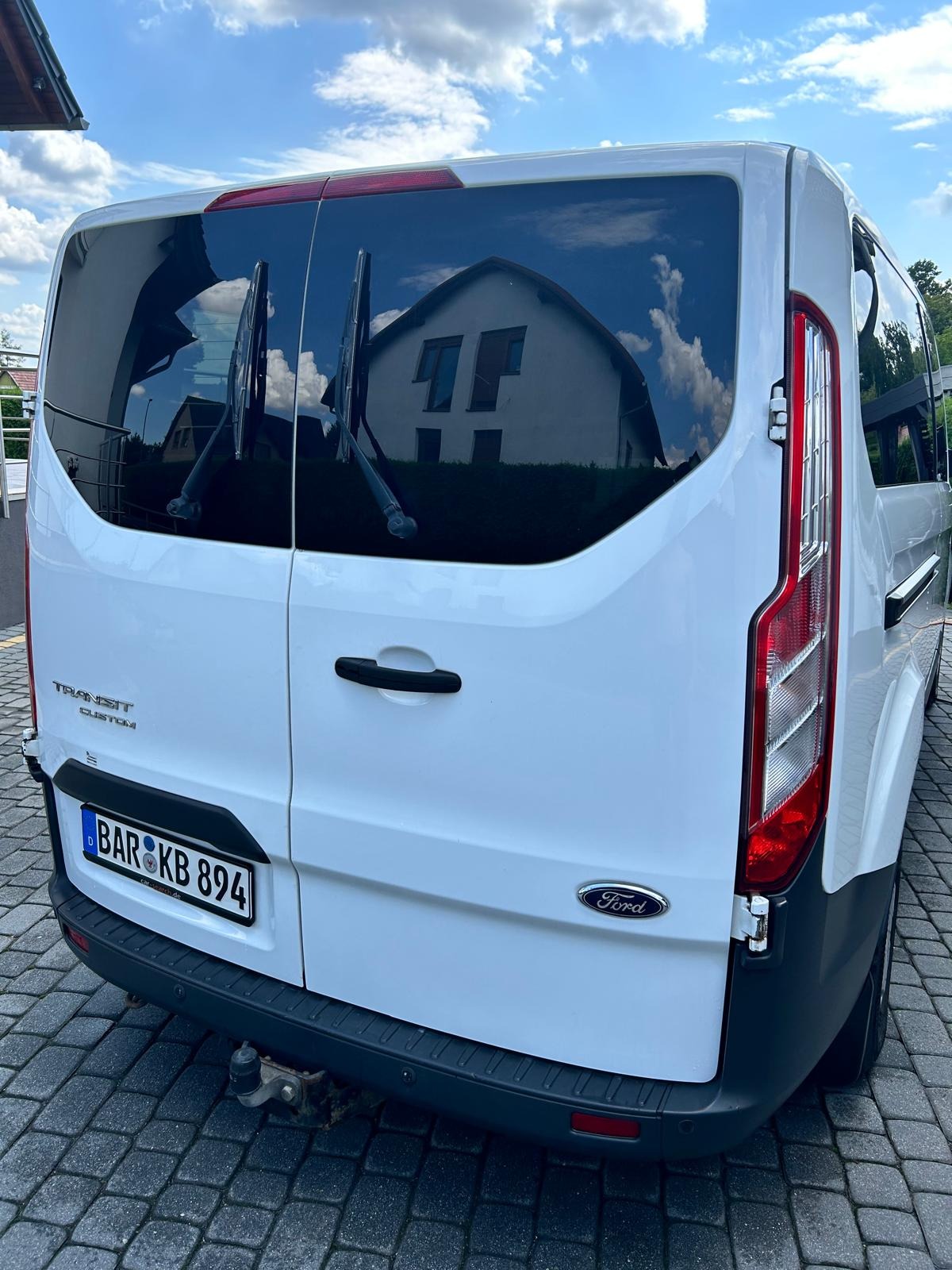 Tył białego Forda Transita Custom z widoczną tablicą rejestracyjną i odbiciem budynku w szybie. Auto stoi na kostce brukowej w słoneczny dzień.