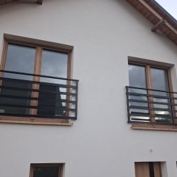 Paweł Otta - Dwa okna z czarnymi, metalowymi balustradami o prostym, nowoczesnym designie na jasnej fasadzie domu. Balustrady zamontowane na drewnianych elementach pod oknami.