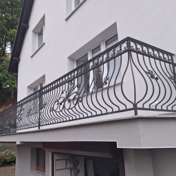 Paweł Otta - Kuta, czarna balustrada balkonowa z dekoracyjnymi elementami roślinnymi, zamontowana na balkonie budynku z białą elewacją. Widok z dołu, zieleń w tle.