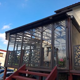 Paweł Otta - Zadaszony taras z ażurowymi panelami i szklanymi ścianami, widok z dołu na tle błękitnego nieba. Ciemnobrązowa konstrukcja z czerwonymi schodami.