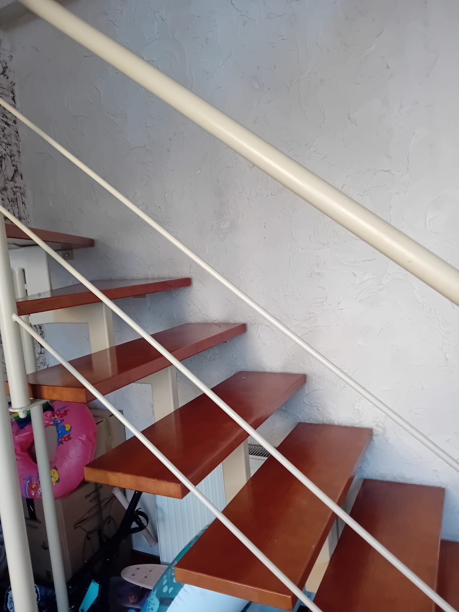 Spiralne schody z drewnianymi stopniami i białą metalową balustradą na tle ściany o nierównej fakturze. Ujęcie od dołu, widoczne przedmioty pod schodami.