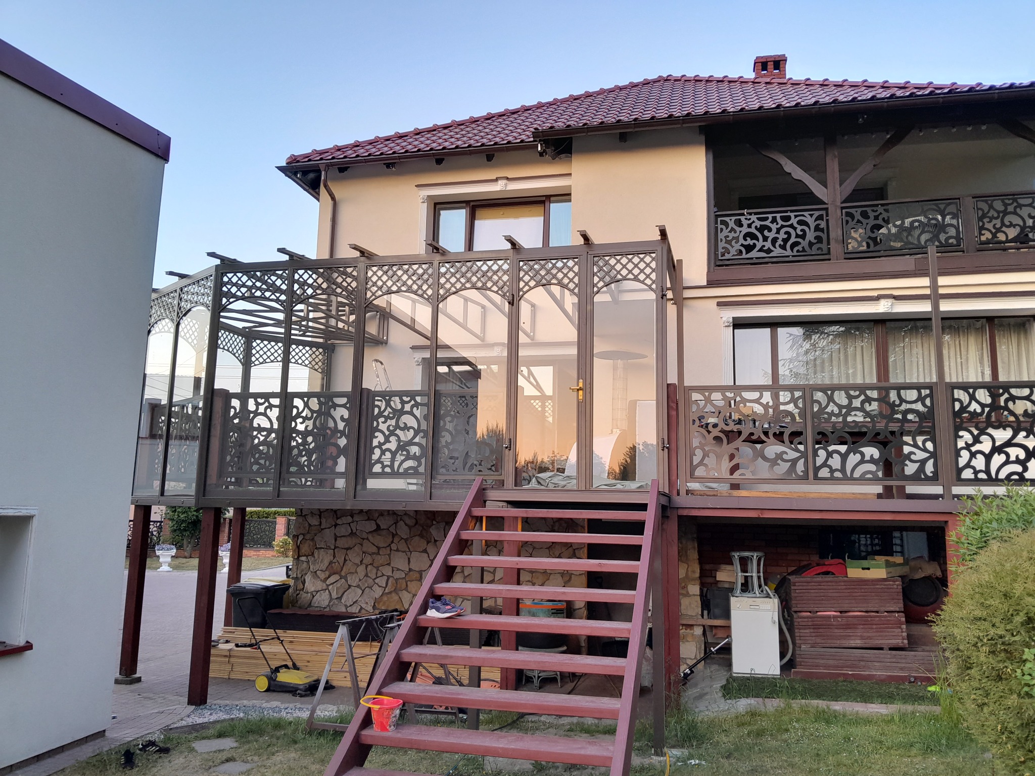 Zabudowa balkonu z ażurowymi panelami i szklanymi elementami, brązowa konstrukcja, widok z dołu na schody wejściowe, dom z balkonem w tle.