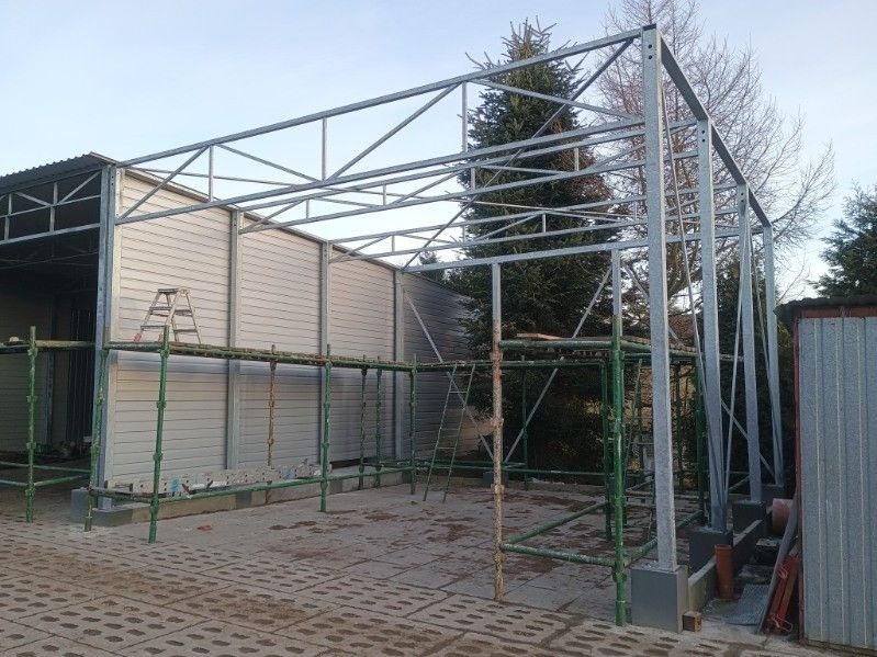 Szkielet metalowej konstrukcji z rusztowaniem na zewnątrz budynku. Widoczne elementy spawane i skręcane. W tle drzewa i fragmenty zabudowań.