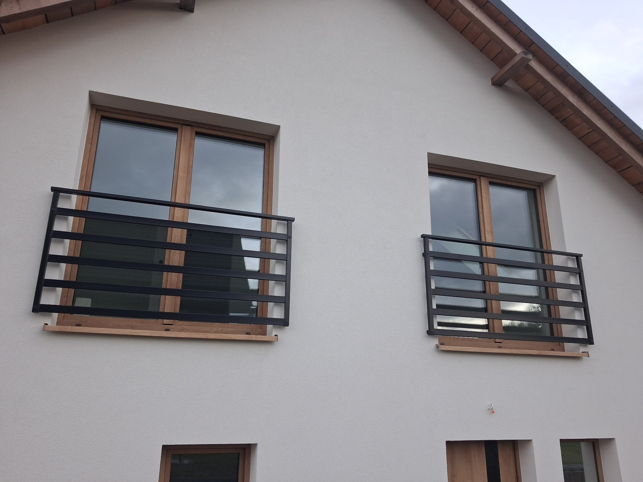 Dwa okna z czarnymi, metalowymi balustradami o prostym, nowoczesnym designie na jasnej fasadzie domu. Balustrady zamontowane na drewnianych elementach pod oknami.