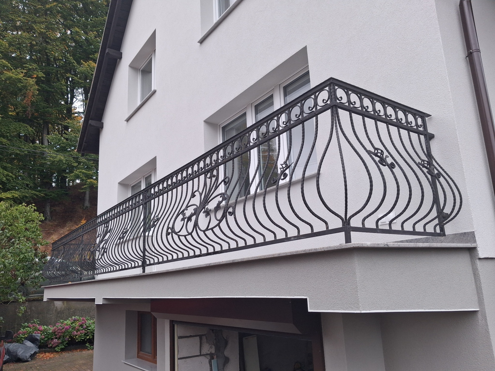 Kuta, czarna balustrada balkonowa z dekoracyjnymi elementami roślinnymi, zamontowana na balkonie budynku z białą elewacją. Widok z dołu, zieleń w tle.