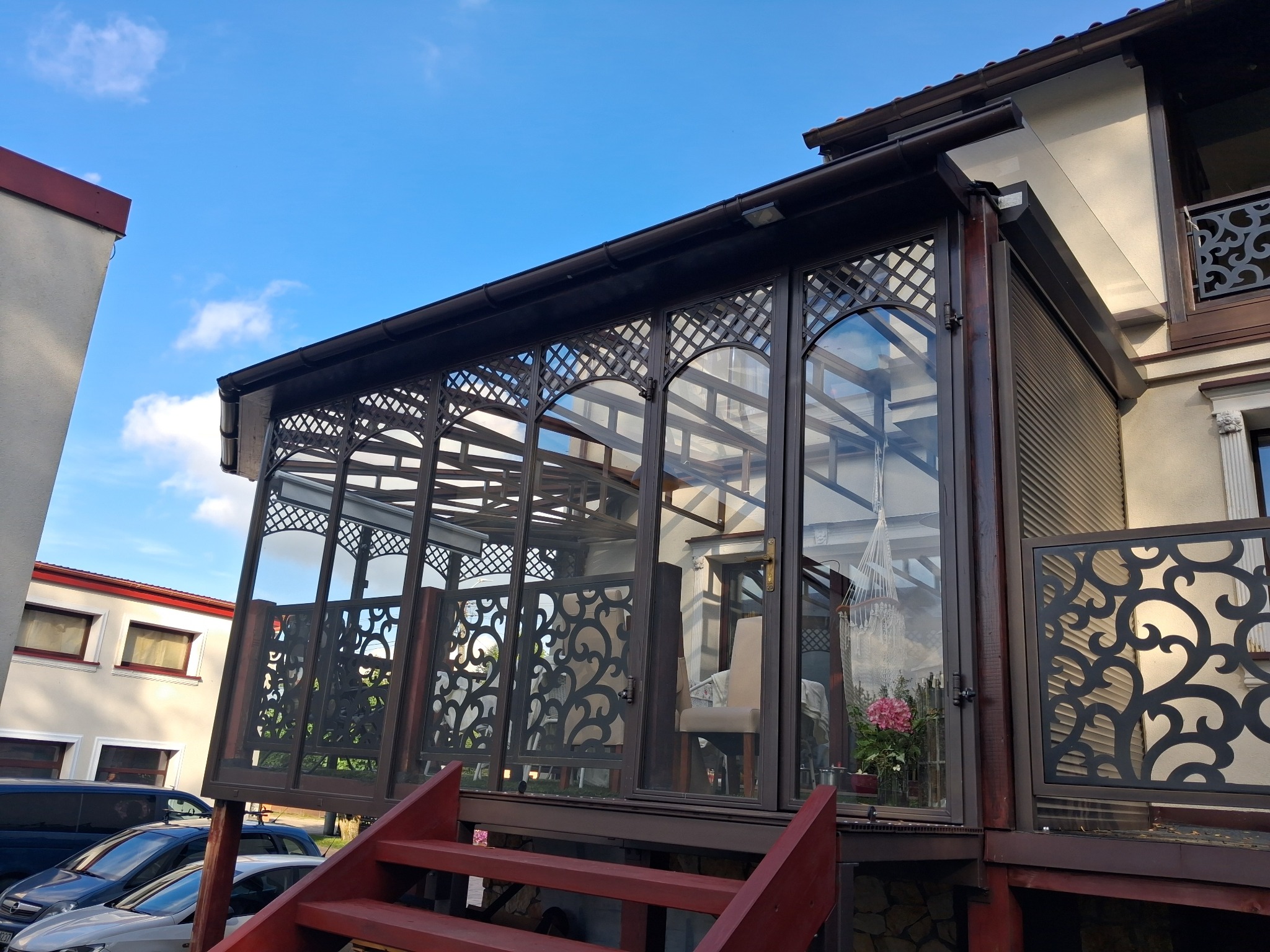 Zadaszony taras z ażurowymi panelami i szklanymi ścianami, widok z dołu na tle błękitnego nieba. Ciemnobrązowa konstrukcja z czerwonymi schodami.