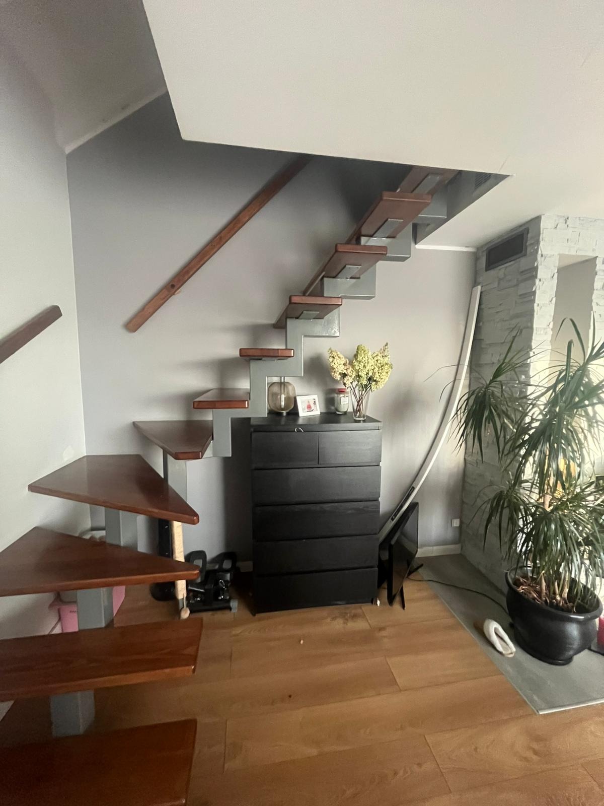 Nowoczesne schody wspornikowe z drewnianymi stopniami i szarą konstrukcją, umieszczone w jasnym wnętrzu z komodą i rośliną doniczkową. Minimalistyczny design.