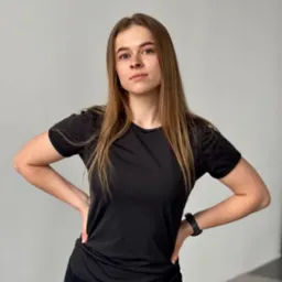 Kobieta w czarnym stroju sportowym, z rękami na biodrach, prezentuje postawę w jasnym wnętrzu, z widocznym zegarkiem sportowym i zieloną matą do ćwiczeń.