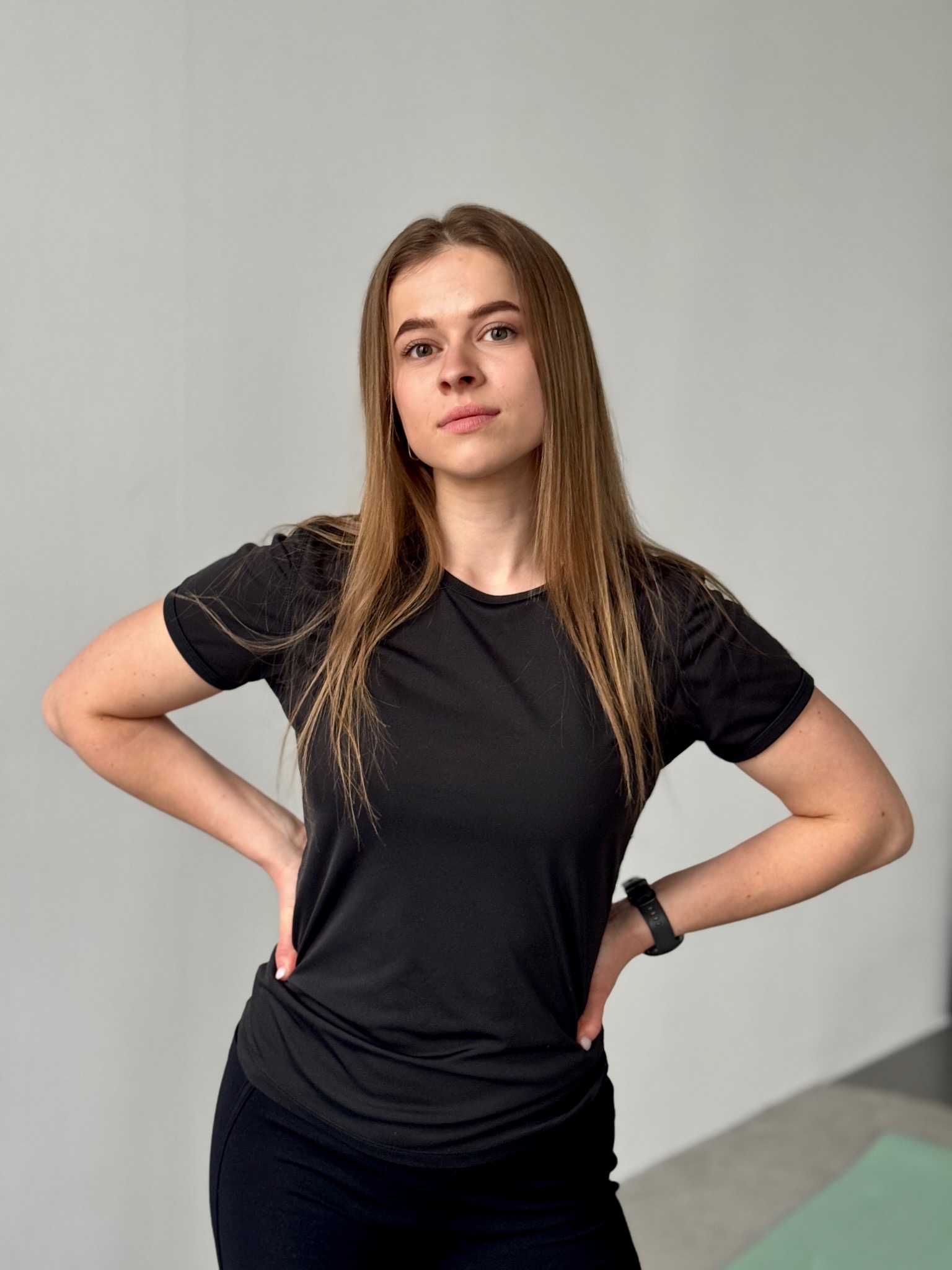 Kobieta w czarnym stroju sportowym, z rękami na biodrach, prezentuje postawę w jasnym wnętrzu, z widocznym zegarkiem sportowym i zieloną matą do ćwiczeń.