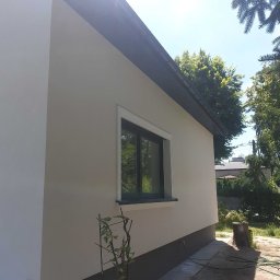 Adam Melon - Jasna elewacja domu jednorodzinnego z ciemną podmurówką i oknem w ciemnej ramie, otoczona zielenią. Widoczny fragment dachu i rynny. Słoneczny dzień.