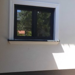 Adam Melon - Elewacja budynku z oknem w czarnej ramie, otoczonym białą dekoracyjną listwą. Widoczna faktura tynku i cień padający na ścianę. Detal wykończenia.