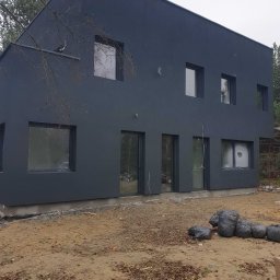 Adam Melon - Nowoczesny, dwukondygnacyjny dom z ciemną elewacją i prostokątnymi oknami. Widoczne worki z odpadami budowlanymi na nierównym terenie.