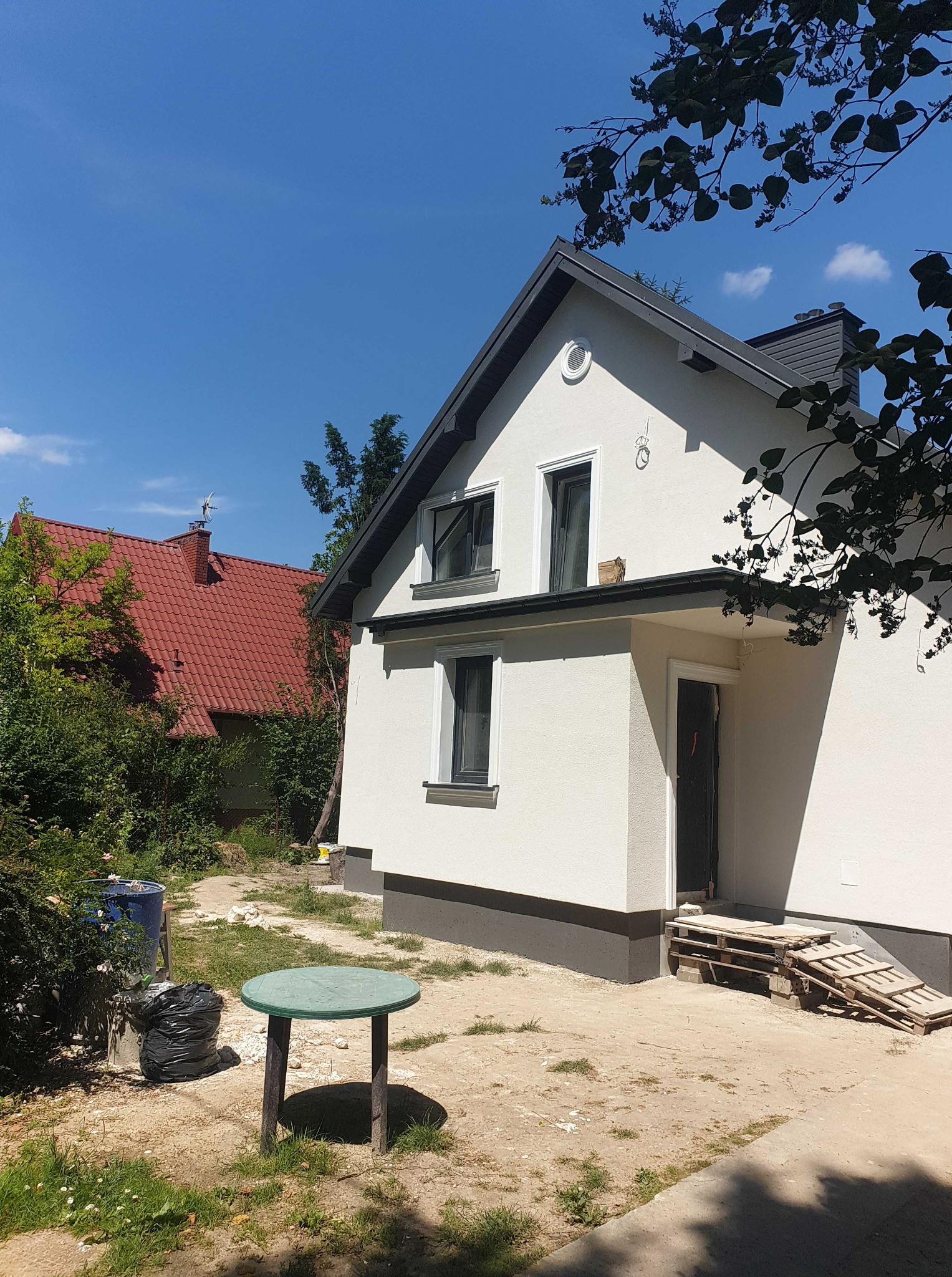 Biała elewacja domu jednorodzinnego z szarymi detalami, widoczna w słoneczny dzień. Na pierwszym planie zielony stolik i zieleń, w tle dom z czerwoną dachówką.