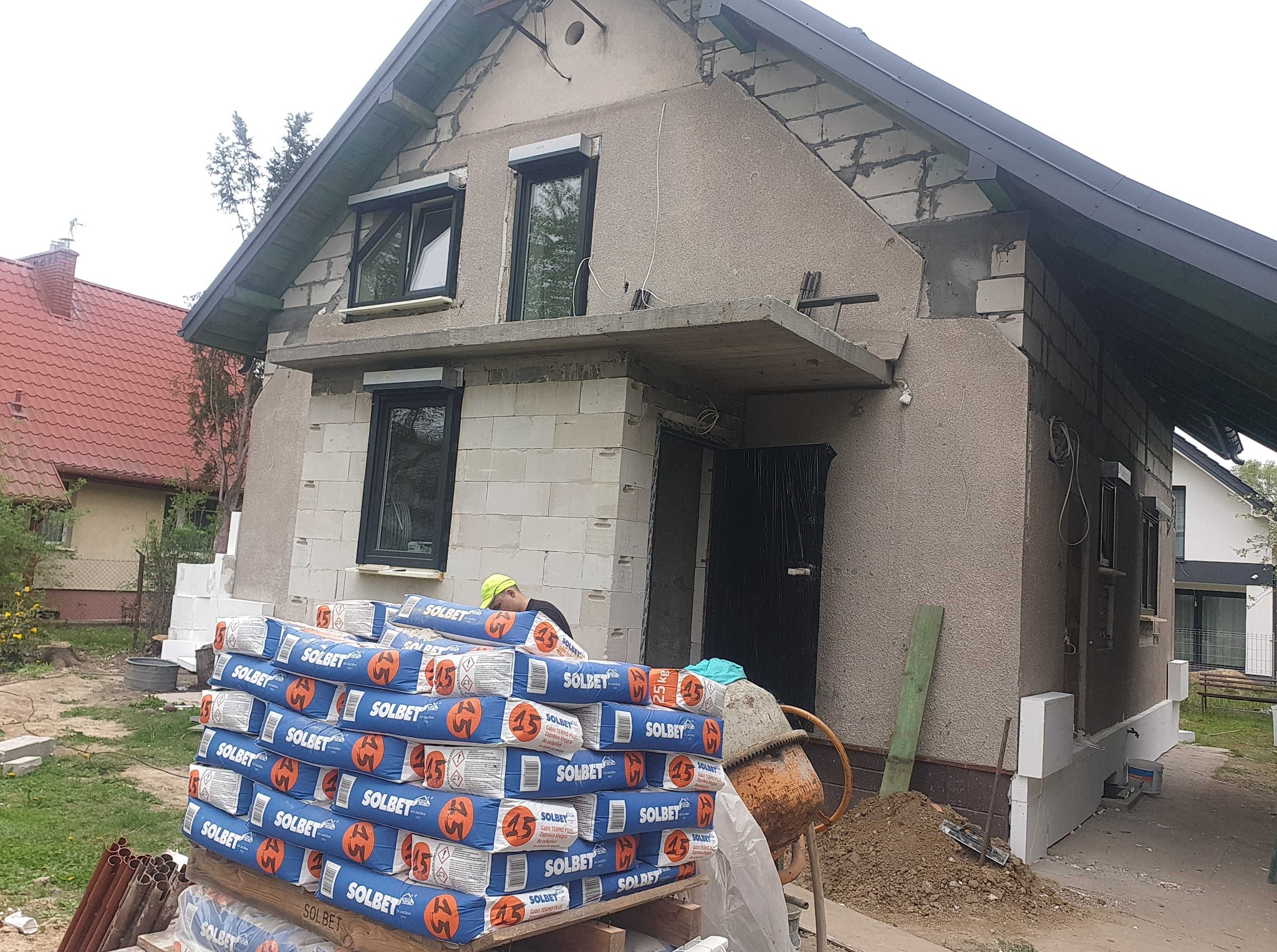 Dom w trakcie ocieplania: widoczne worki z zaprawą Solbet, częściowo otynkowana elewacja i bloczki. Mieszarka betonu obok. Prace elewacyjne w toku.