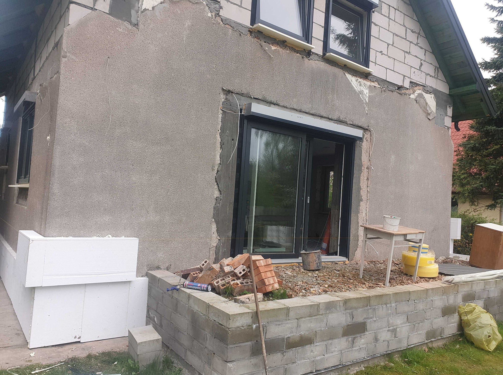 Dom w trakcie ocieplania elewacji: szary tynk, białe płyty styropianowe, cegły, okno i stół z narzędziami. Widoczne niedokończone prace.