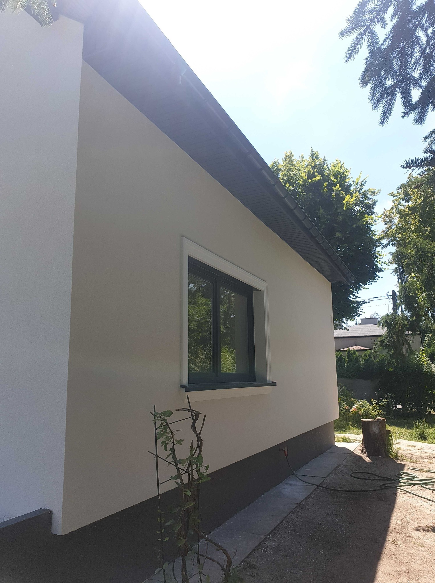 Jasna elewacja domu jednorodzinnego z ciemną podmurówką i oknem w ciemnej ramie, otoczona zielenią. Widoczny fragment dachu i rynny. Słoneczny dzień.