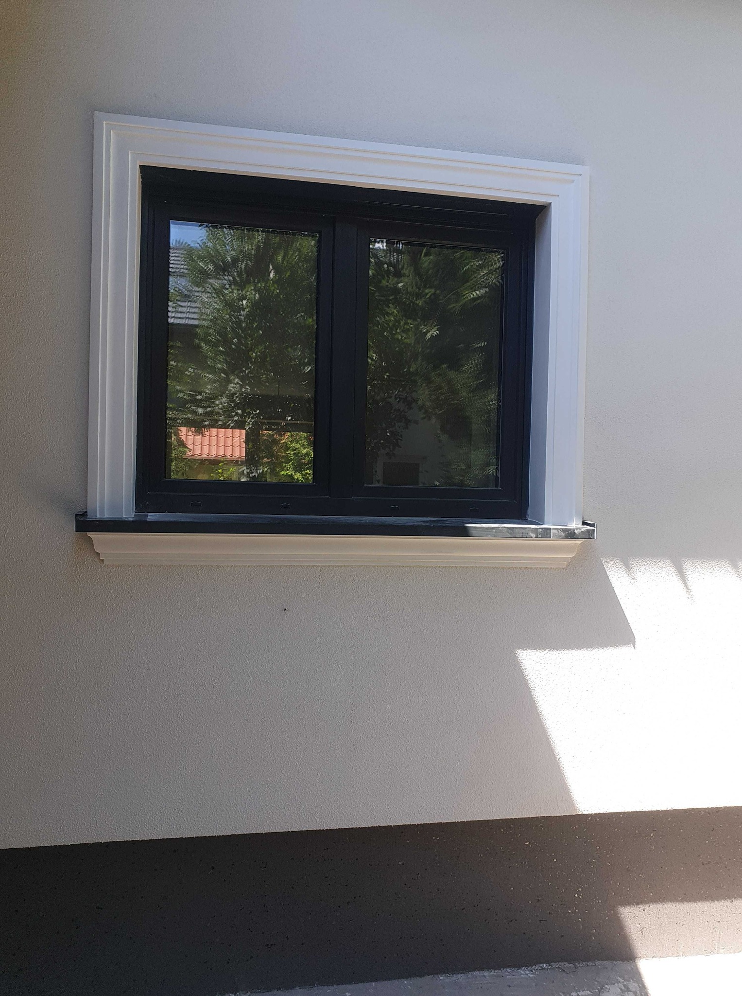 Elewacja budynku z oknem w czarnej ramie, otoczonym białą dekoracyjną listwą. Widoczna faktura tynku i cień padający na ścianę. Detal wykończenia.