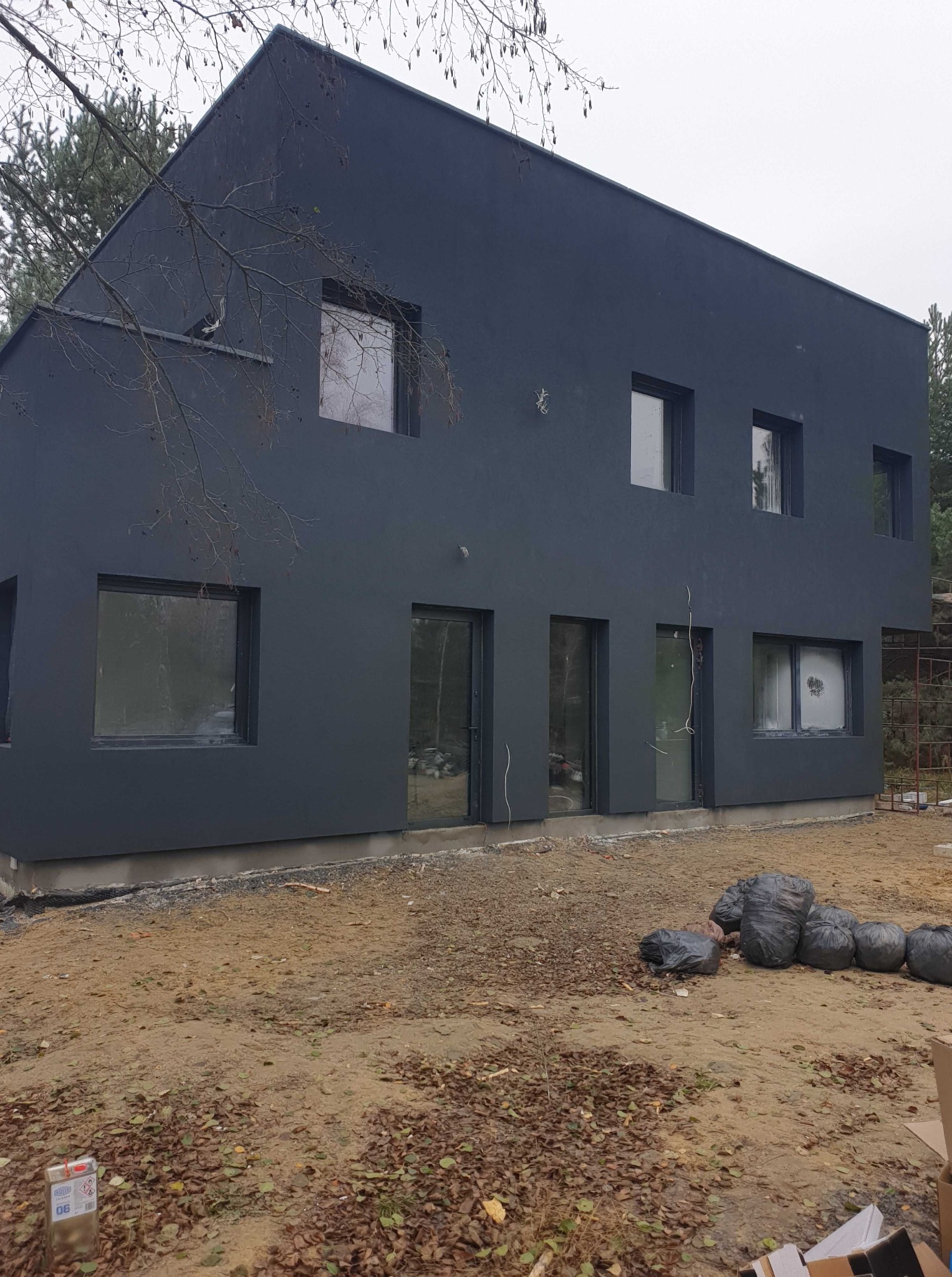 Nowoczesny, dwukondygnacyjny dom z ciemną elewacją i prostokątnymi oknami. Widoczne worki z odpadami budowlanymi na nierównym terenie.