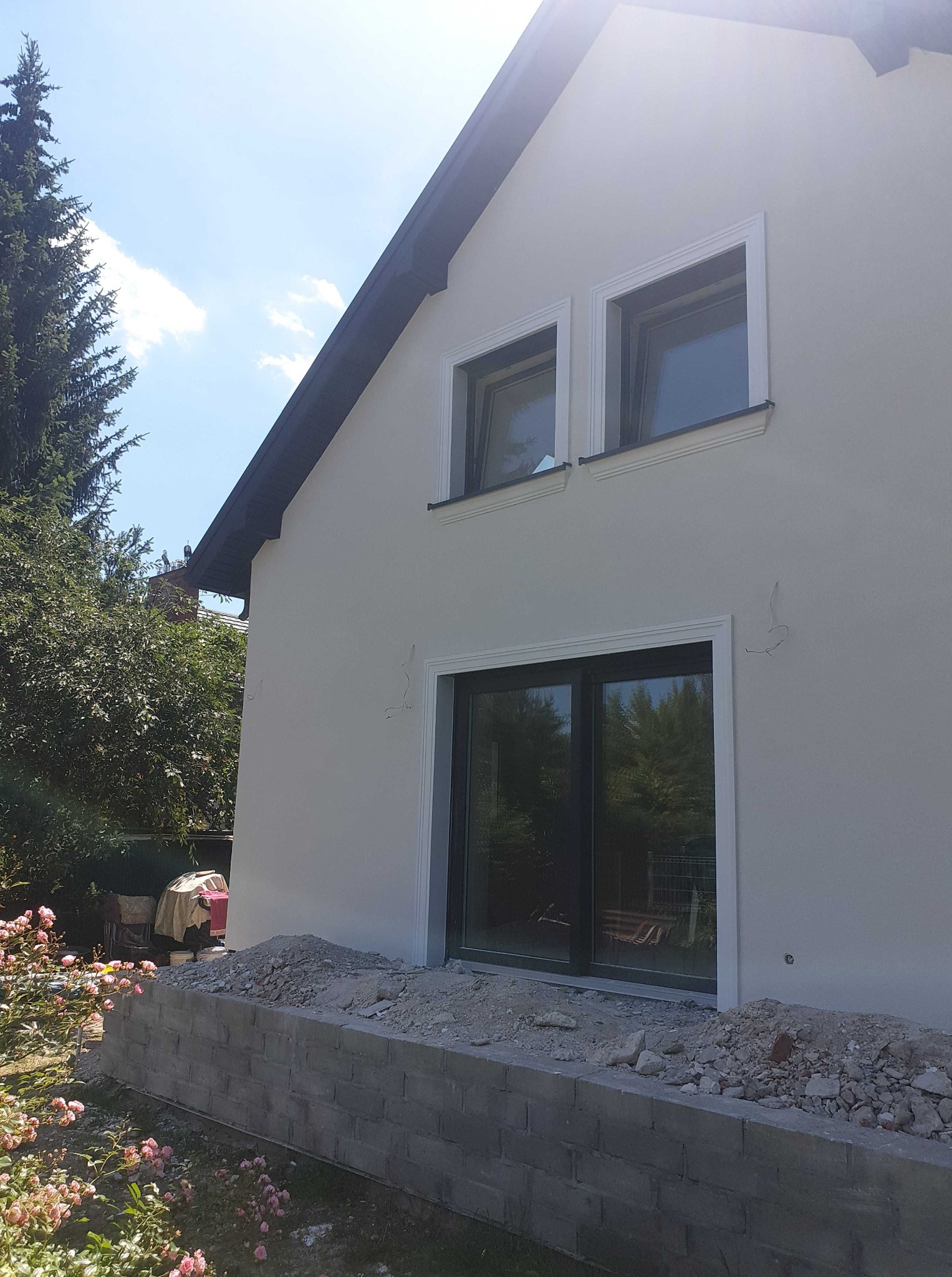 Ściana budynku po tynkowaniu z dekoracyjnymi obramowaniami okiennymi, widoczne ślady po mocowaniu rusztowania, gruz przy fundamencie, róże w ogrodzie.