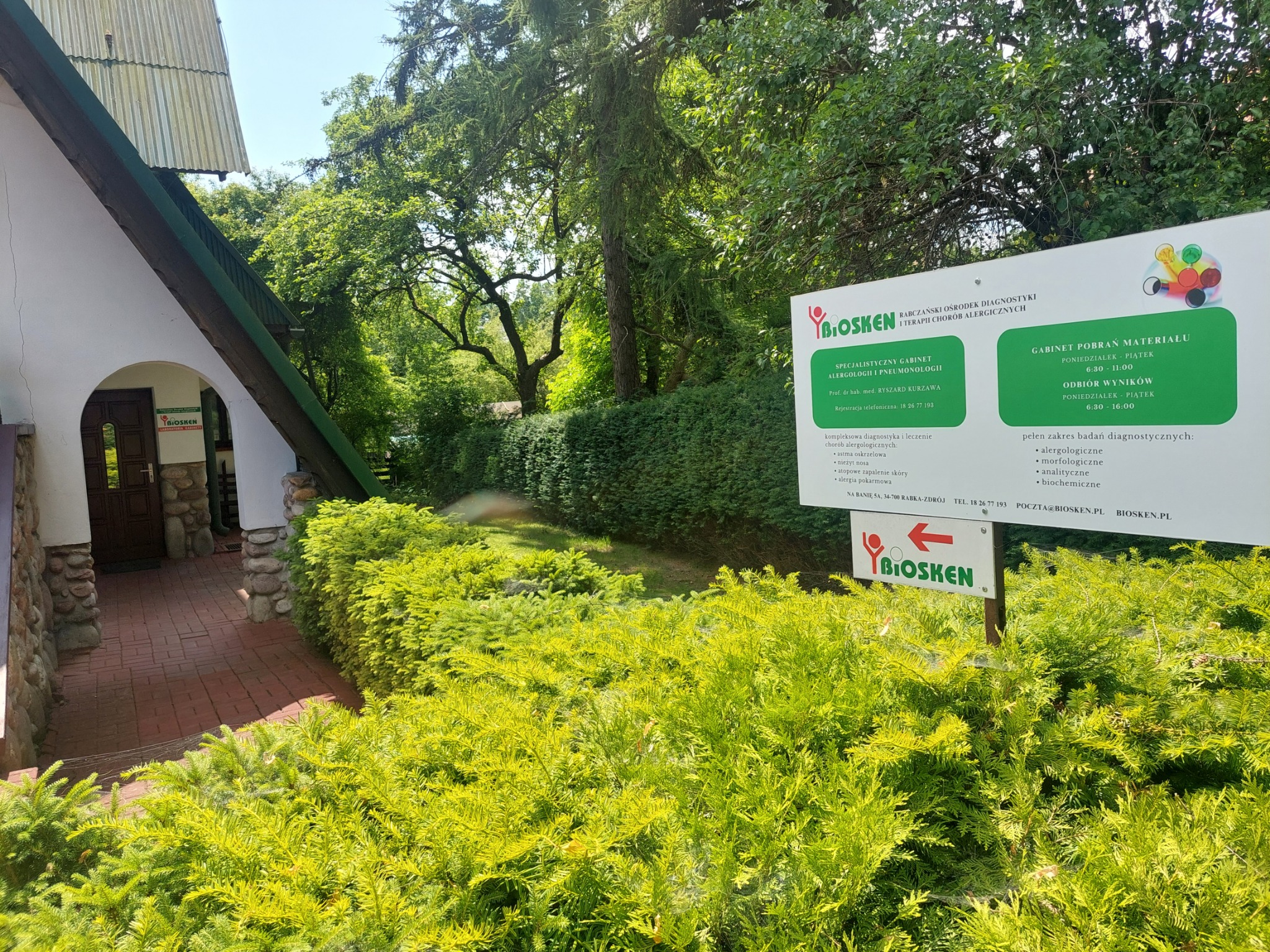 Wejście do gabinetu alergologicznego z tablicą informacyjną o godzinach pracy i zakresie badań, otoczone zielenią. Widok w słoneczny dzień.