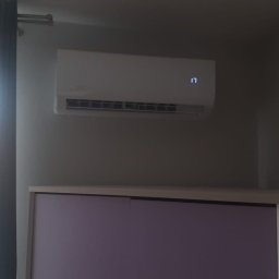 Chłodna Klima - Biała klimatyzacja ścienna w pokoju, nad fioletowymi szafkami, z wyświetlaczem ustawionym na 17 stopni, obok okna z zasłoną.