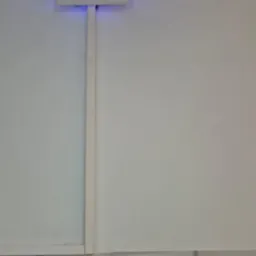 Biały kwadratowy panel sterowania z niebieską diodą LED na białej ścianie, z widoczną listwą przypodłogową i dekoracyjną fakturą ściany poniżej.