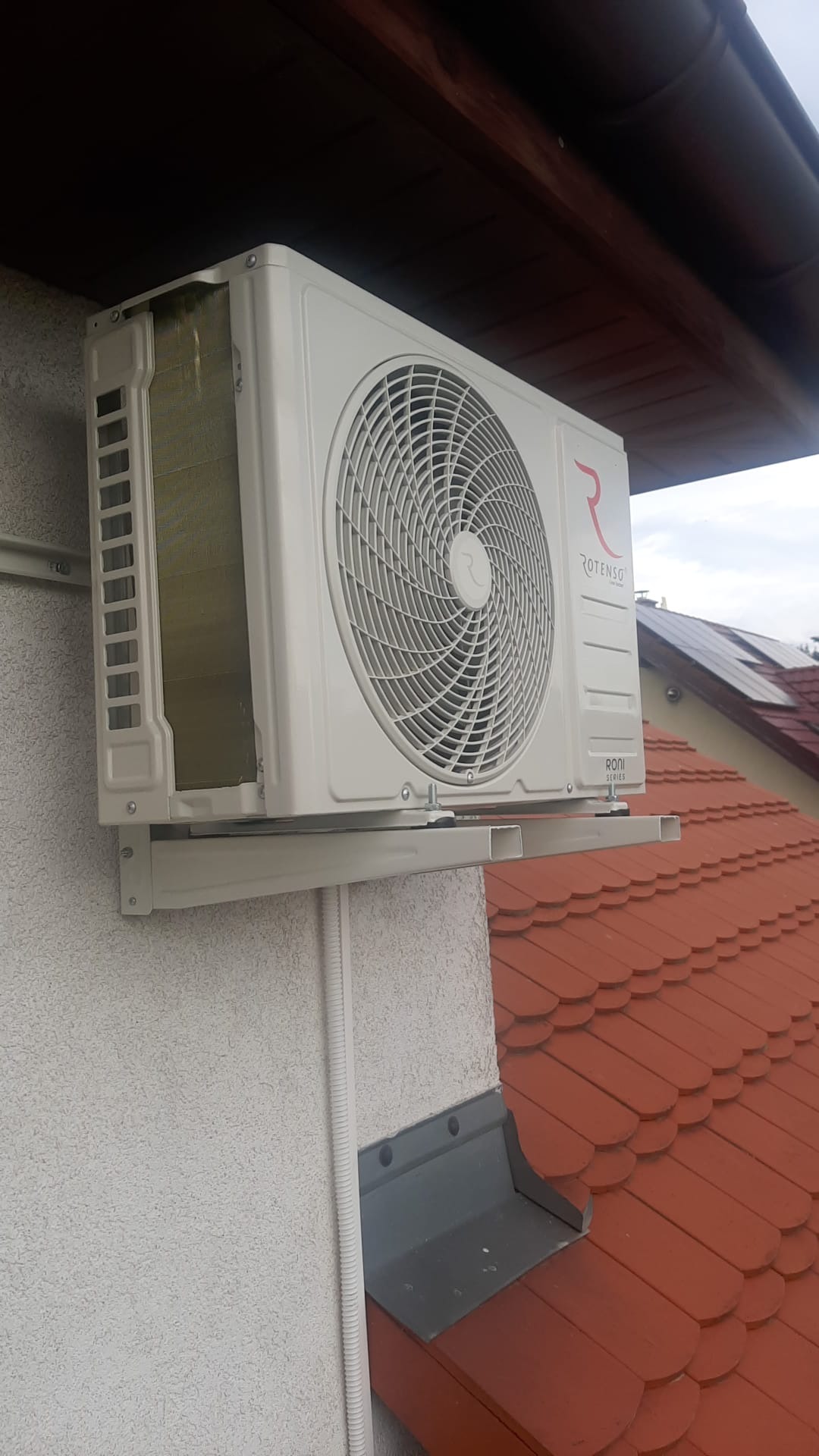 Klimatyzator Rotenso Roni Series zamontowany na elewacji budynku z czerwonym dachem. Widoczne rury instalacyjne i wspornik montażowy.