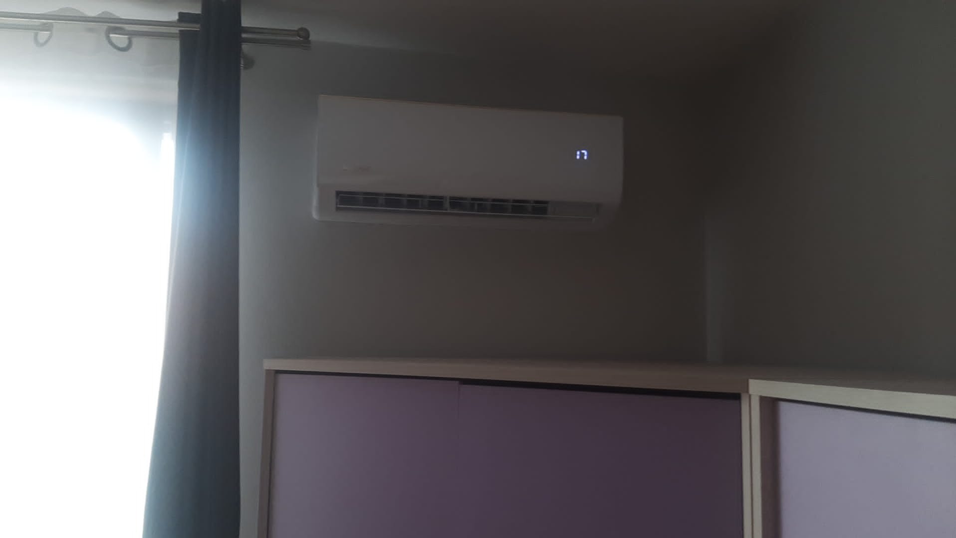 Biała klimatyzacja ścienna w pokoju, nad fioletowymi szafkami, z wyświetlaczem ustawionym na 17 stopni, obok okna z zasłoną.