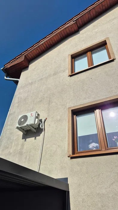 Boczny widok domu z zamontowaną klimatyzacją na elewacji, widoczne okna z brązowymi ramami i fragment dachu z brązową podbitką, błękitne niebo w tle.
