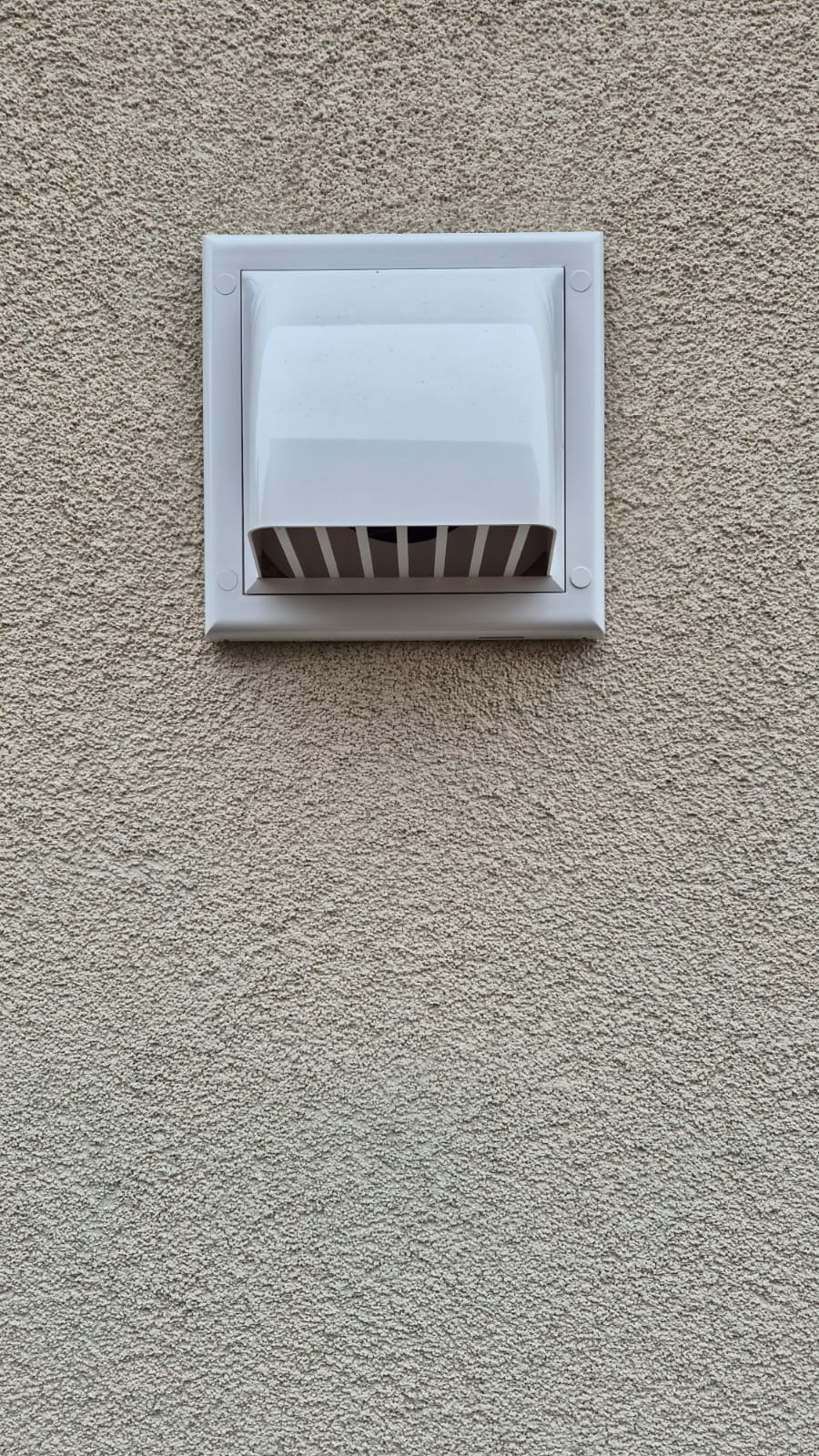 Zamontowany na elewacji budynku biały, prostokątny nawiewnik wentylacyjny z poziomymi kratkami. Widoczna faktura tynku.