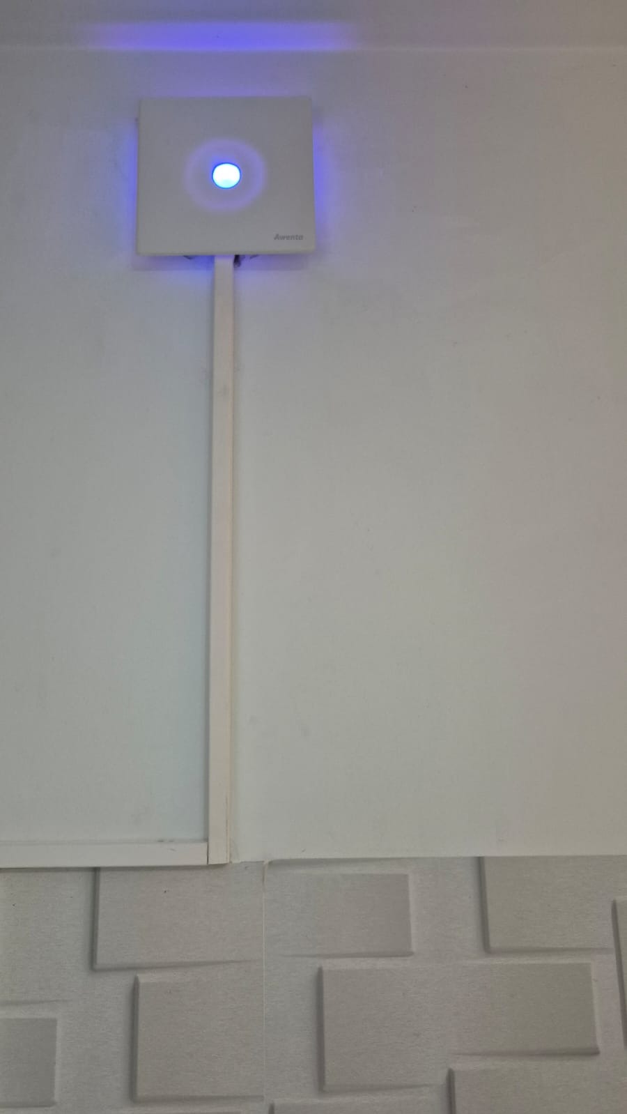 Biały kwadratowy panel sterowania z niebieską diodą LED na białej ścianie, z widoczną listwą przypodłogową i dekoracyjną fakturą ściany poniżej.