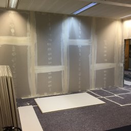 Damian Paszenda - Wnętrze w trakcie prac z płytami gipsowo-kartonowymi: ściany z widocznymi łączeniami, płyty na podłodze, przygotowane do montażu w pomieszczeniu biurowym.